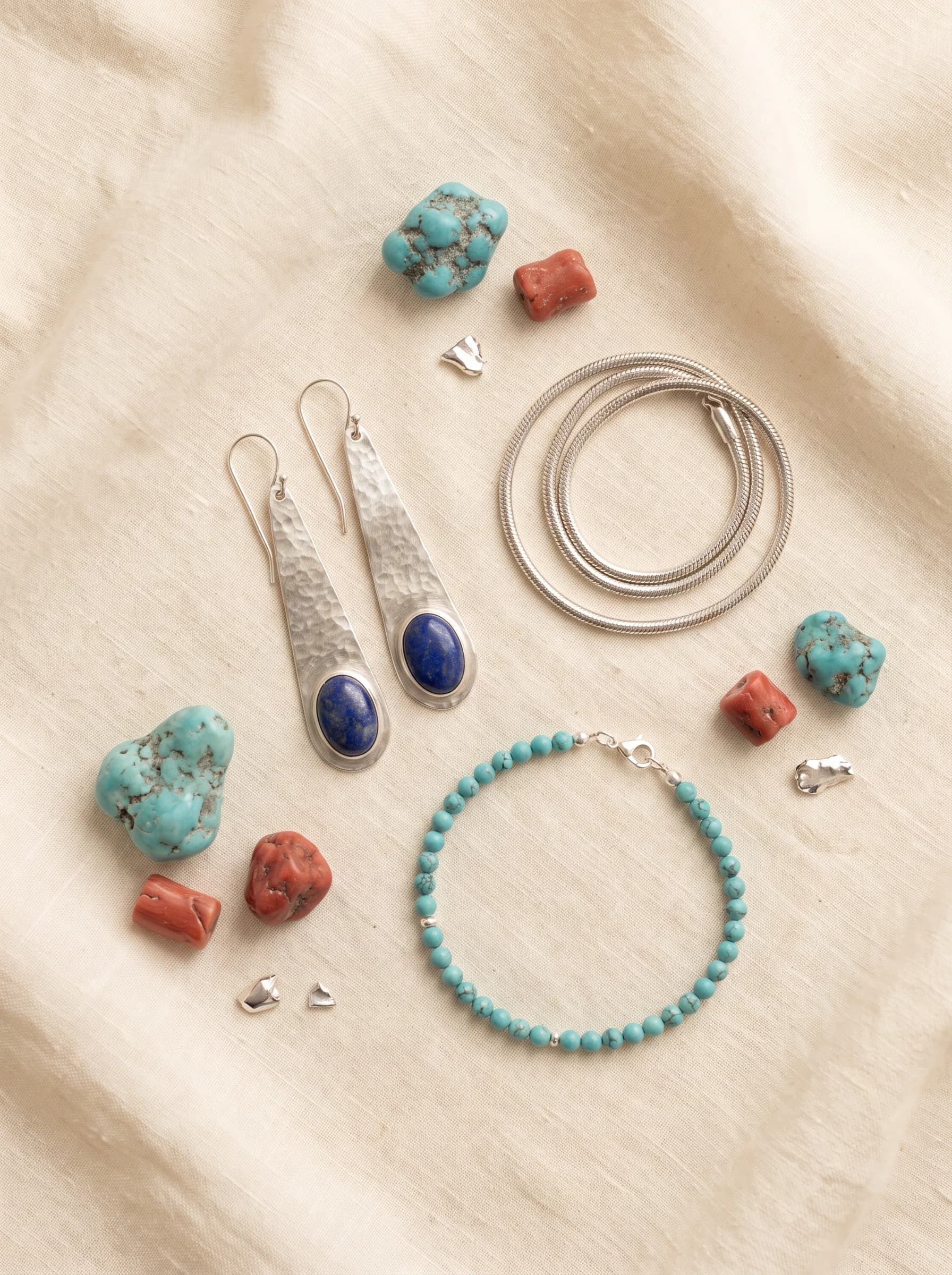 Flat lay of silver drop earrings with lapis lazuli, a snake chain and a turquoise bead bracelet on cream linen