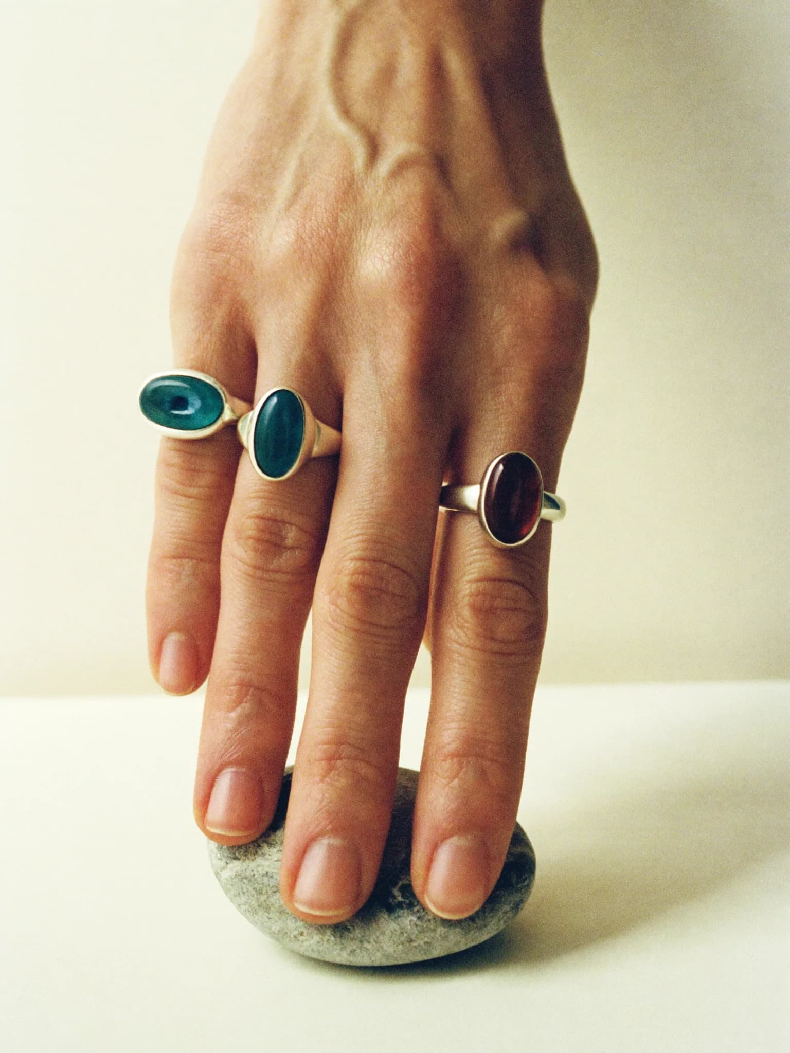 Hand wearing three sterling silver rings with turquoise and garnet stones, fingertips resting on a grey pebble